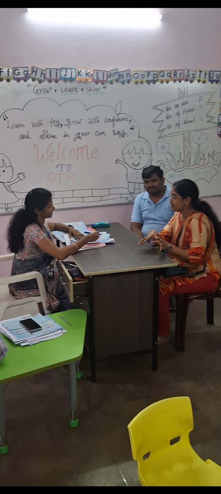 Three people sit at a classroom table, discussing documents, with colorful wall charts and alphabet decorations behind.